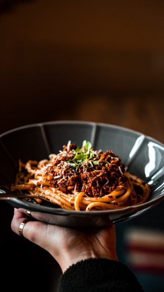 Holding blue bowl of Wild Game Bolognese topped with parmesan and basil over spaghetti noodles. 