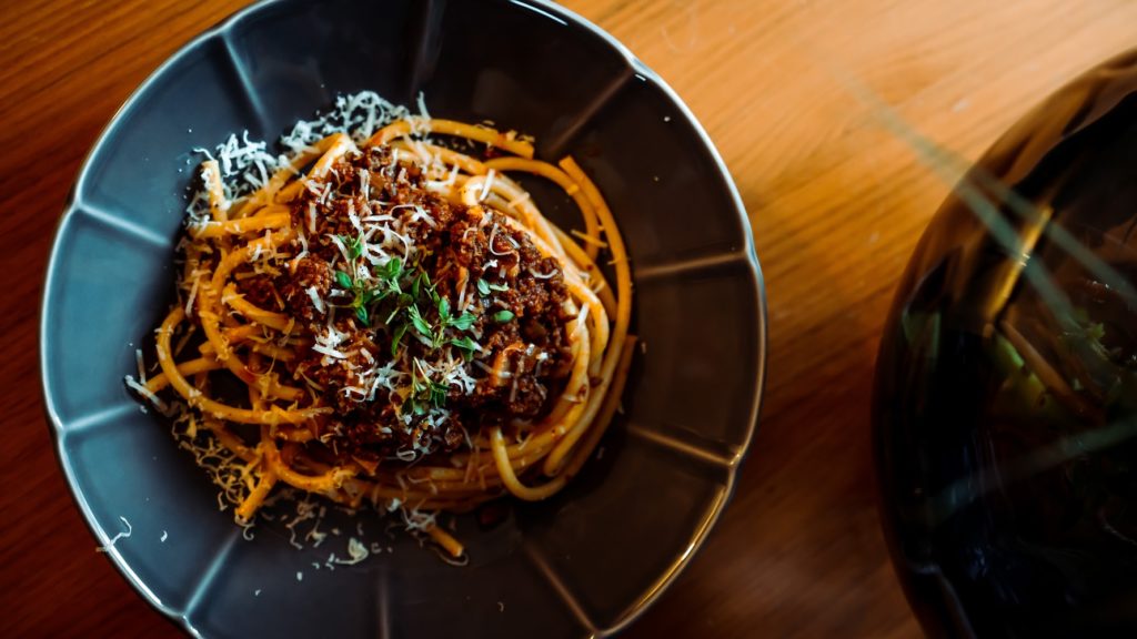 Blue bowl of Wild Game Bolognese topped with parmesan and basil over spaghetti noodles on wood table. 