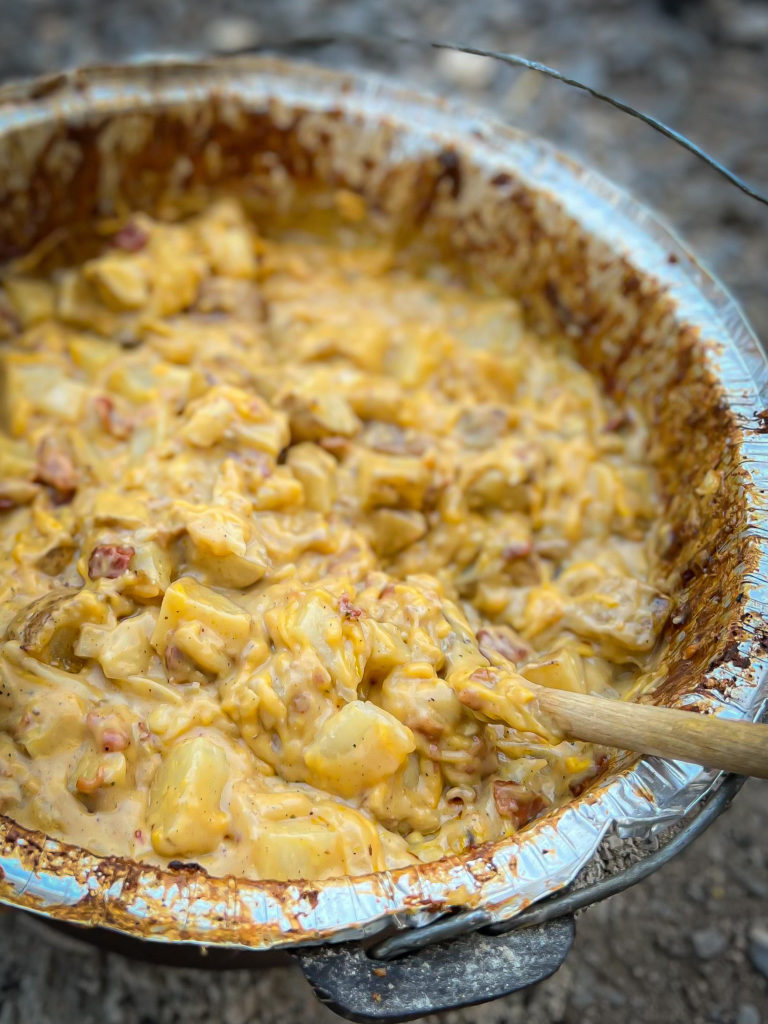 Easy creamy & cheesy dutch oven potatoes with bacon in dutch oven cooked and ready to serve with wooden spoon.